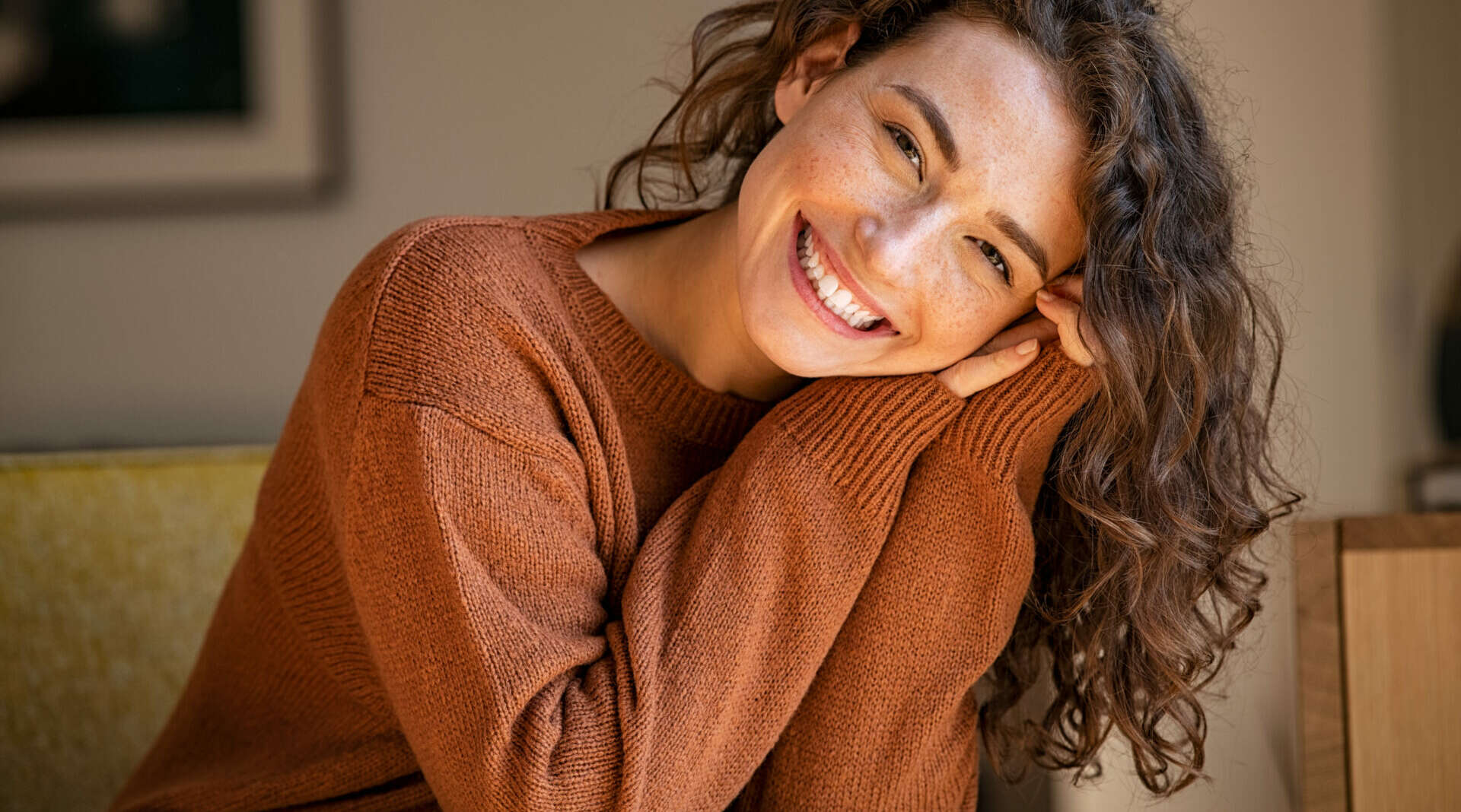 Happy young woman sitting on sofa at home and looking at camera. Portrait of comfortable woman in winter clothes relaxing on armchair. Portrait of beautiful girl smiling and relaxing during autumn.